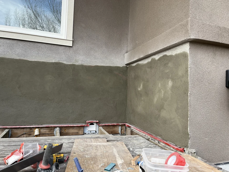 Stucco repair in progress around an exterior window and corner, with fresh cementitious coating and site materials visible.